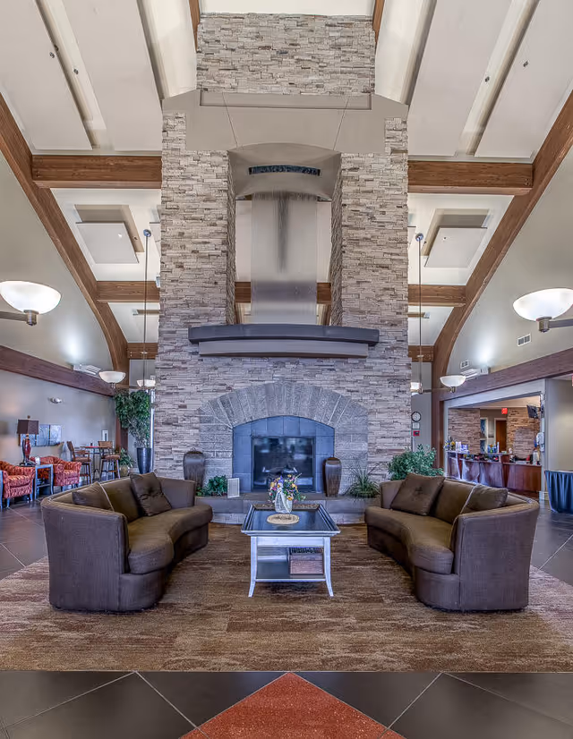 Spacious assisted living lobby with a large stone fireplace, two curved sofas and a coffee table beneath a high beamed ceiling.
