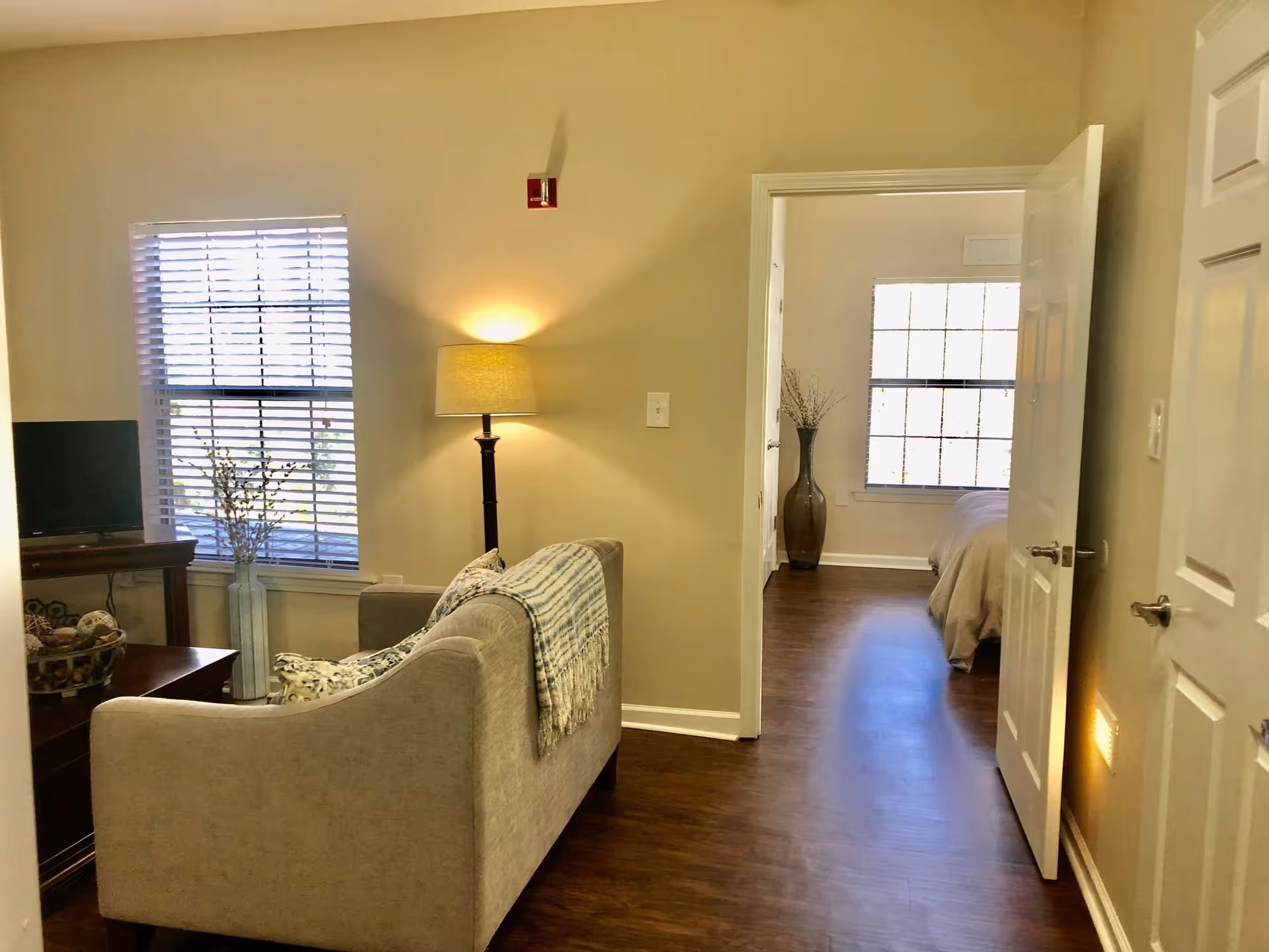 Interior view of a senior living facility showing a cozy living room with a beige sofa, a floor lamp, a window with blinds, and a small TV on a wooden table. An open door leads to a bedroom with a bed and a large window, and a tall vase with decorative branches is placed near the window.