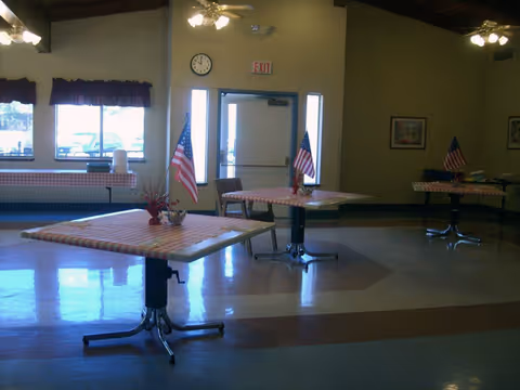 Multipurpose dining room with tables covered in red-checkered tablecloths and small American flags, windows and an exit door in the background.