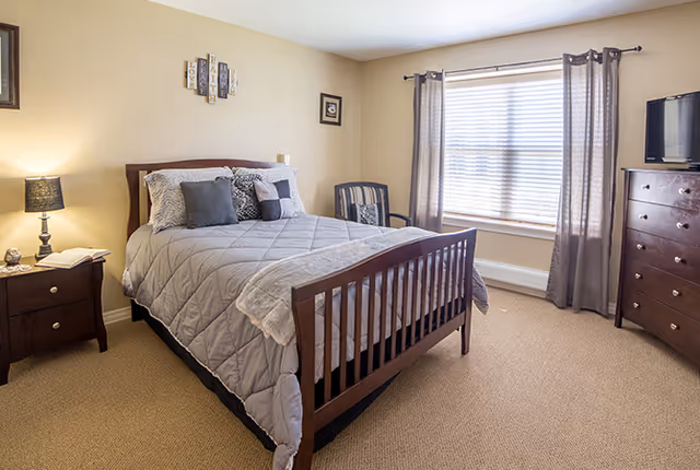 Well-lit bedroom with a wooden bed, nightstand, dresser, armchair, and a large window with curtains.