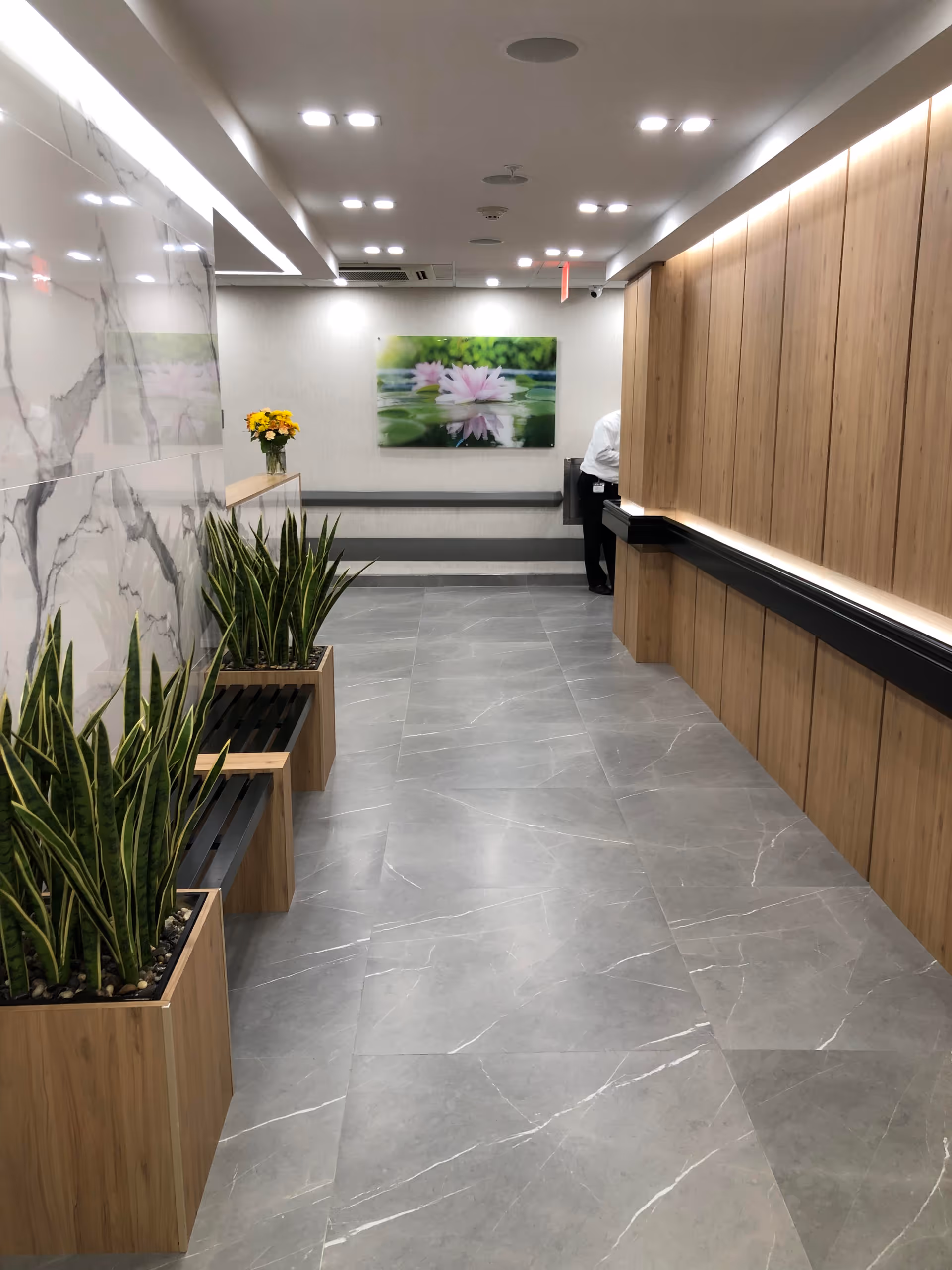 Modern interior hallway with gray tile floor, wood-paneled wall and counter, planters with snake plants, and floral wall art at the far end.