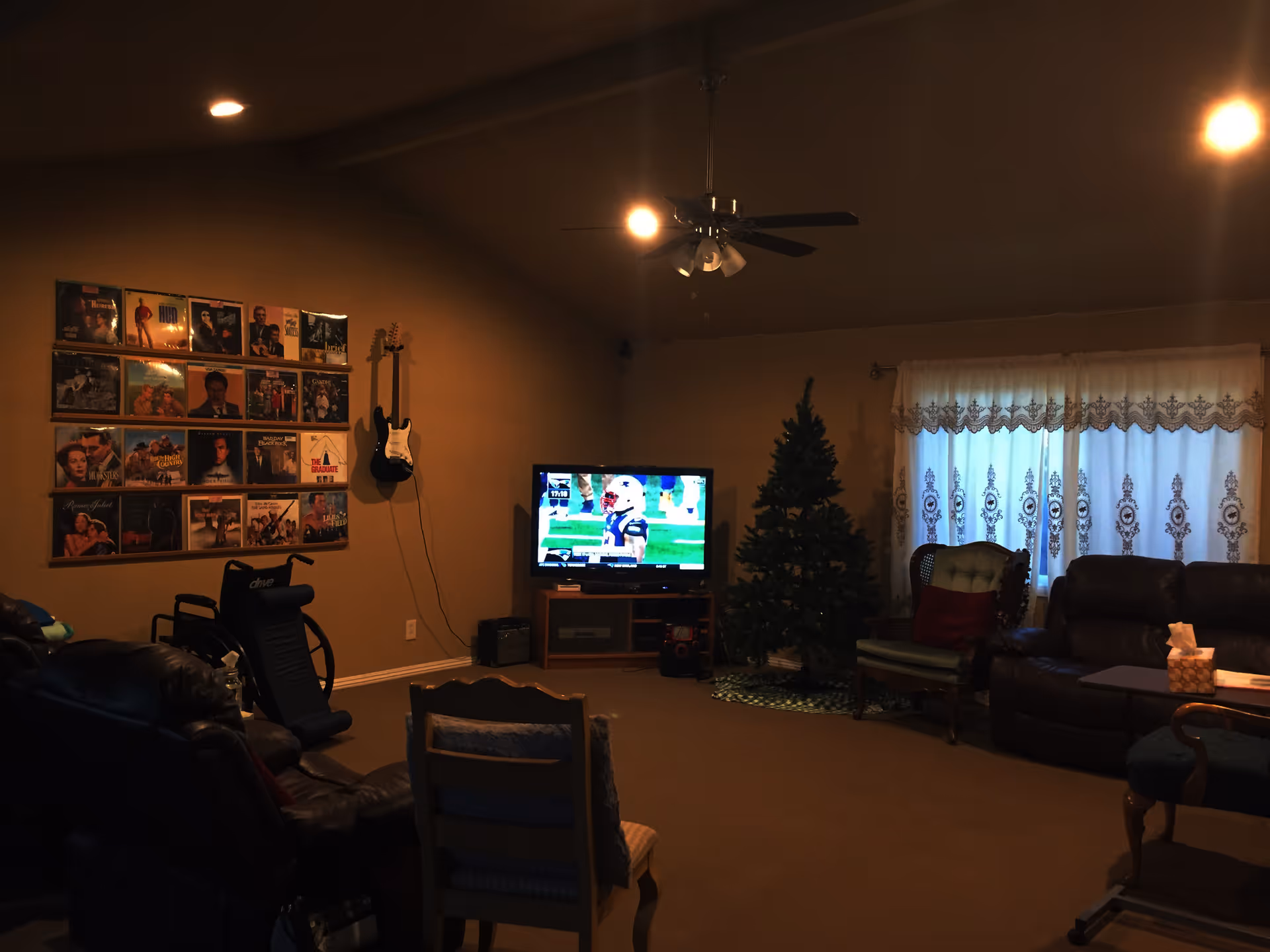 A cozy living room with a television showing a football game, a decorated Christmas tree, a wall-mounted electric guitar, a collection of framed pictures or album covers on the wall, several chairs and a sofa, and a window with lace curtains.