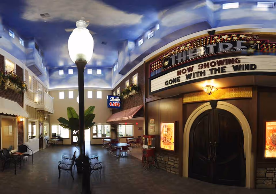 Indoor common area designed to resemble a small town street with a vintage theater entrance showing 'Gone With The Wind', a Pepsi Cafe sign, benches, tables, chairs, and a tall street lamp in the center under a ceiling painted to look like a sky with clouds.
