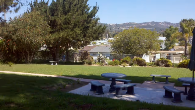 Outdoor area with green grass, trees, and a concrete patio featuring round tables with attached benches. Residential buildings and hills are visible in the background under a clear sky.