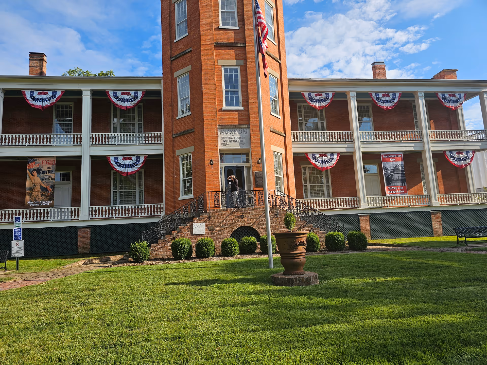 Red brick two-story historic building with wraparound porches decorated with patriotic bunting, a flagpole and manicured lawn in front.