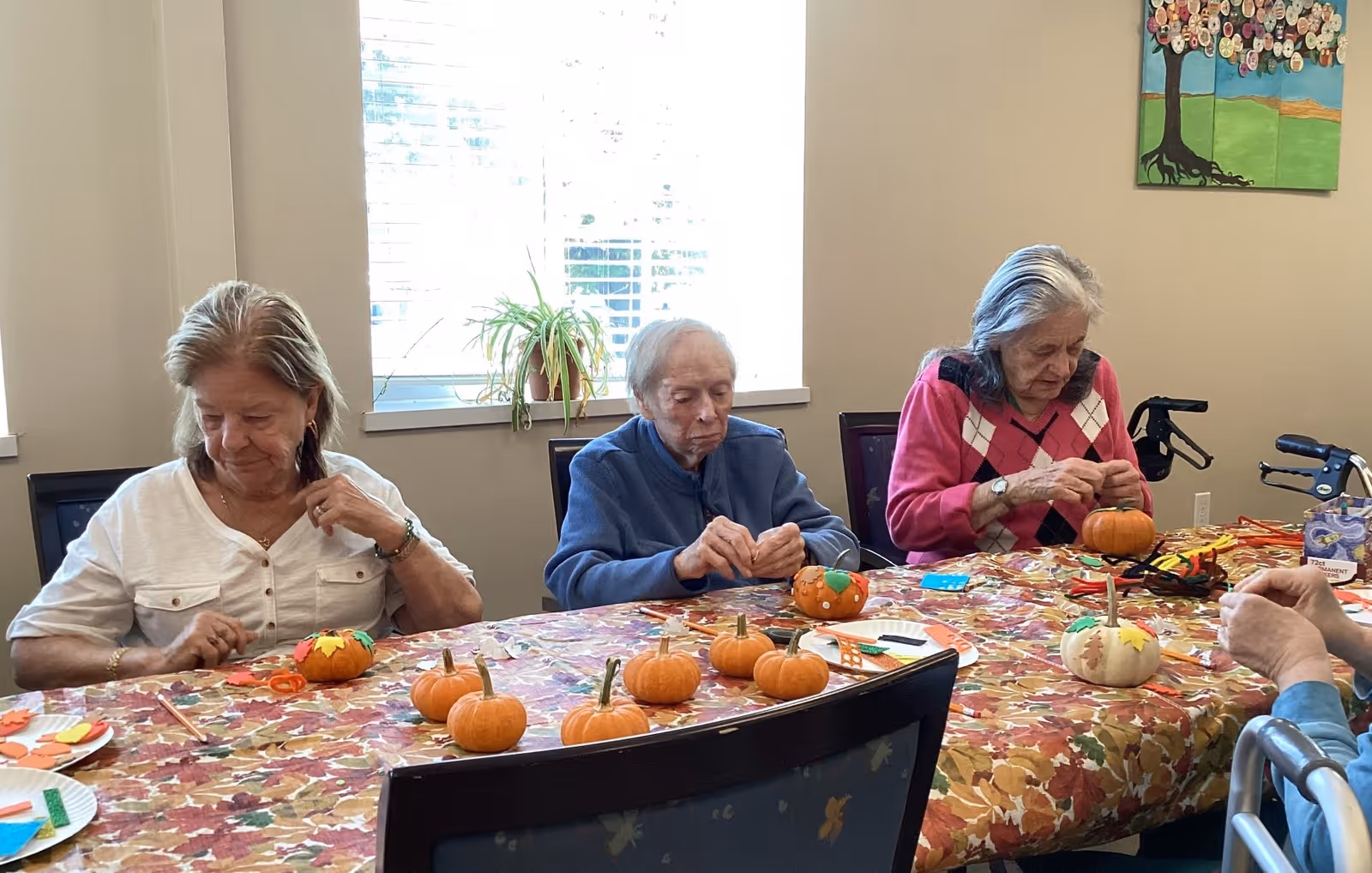 Three elderly women sitting at a table covered with a fall-themed tablecloth, engaging in a craft activity with small pumpkins and colorful materials. A window with blinds and a potted plant is in the background, along with a wall decoration featuring a tree with buttons.