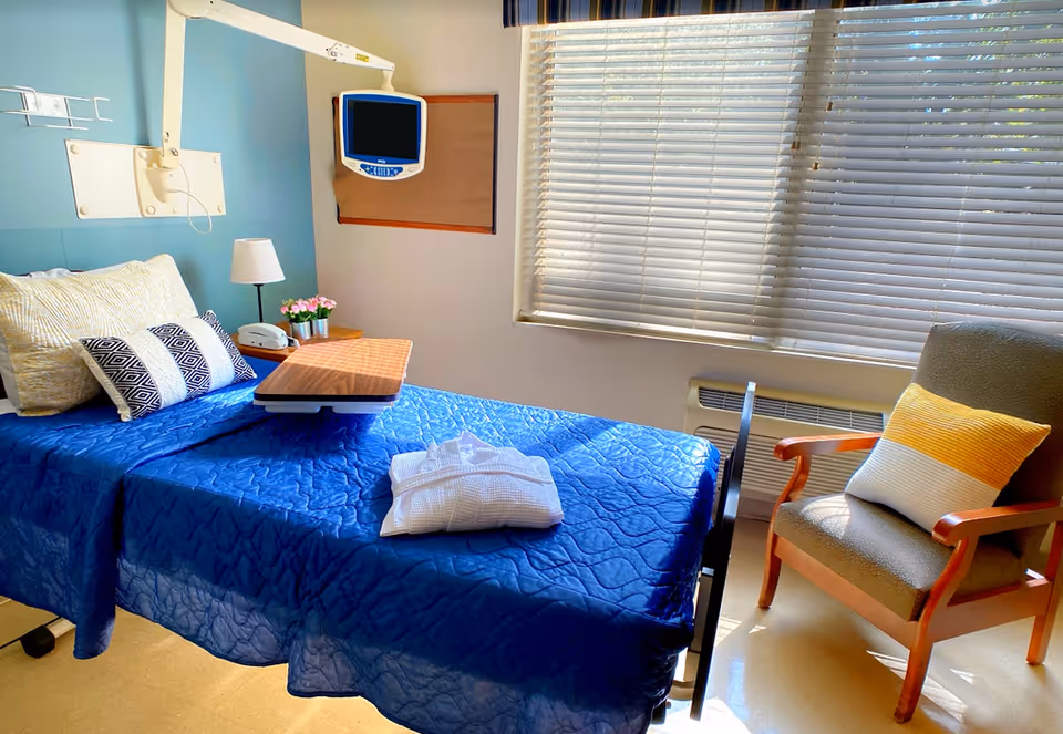 Sunlit patient bedroom in a rehab facility with a blue-covered bed, overbed table, wall-mounted monitor, and an armchair by the window.