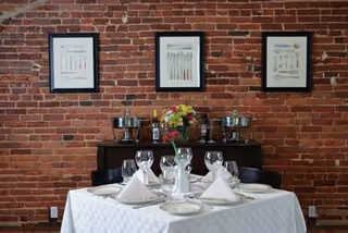 A dining table set with folded napkins, wine glasses and a floral centerpiece in front of a brick wall with framed artwork and a sideboard.