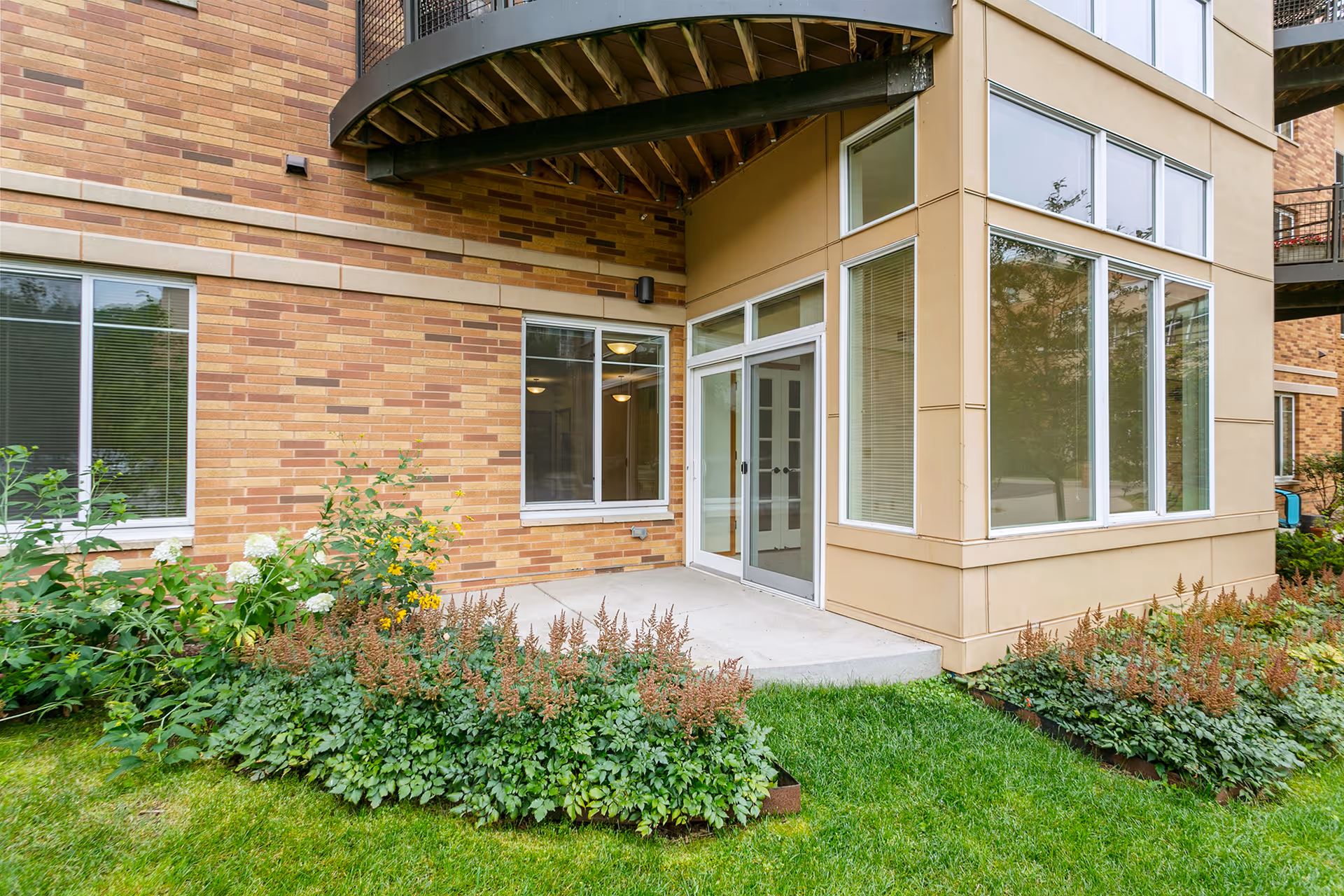 Exterior view of a building corner with large windows and a glass door leading to a small concrete patio surrounded by green grass and flower beds. The building has a brick and beige panel facade with a balcony overhead.