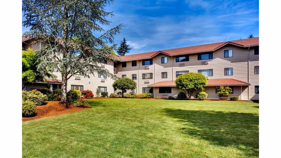 A large, well-maintained grassy courtyard surrounded by a three-story residential building with beige siding and red-tiled roofs. The courtyard features several trees, shrubs, and landscaped garden beds under a partly cloudy blue sky.