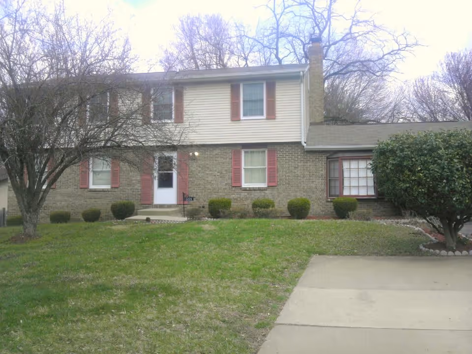Front exterior view of a two-story house with brick and siding walls, red shutters on the windows, a white front door, a chimney, and a driveway. There are leafless trees and trimmed bushes in the front yard.