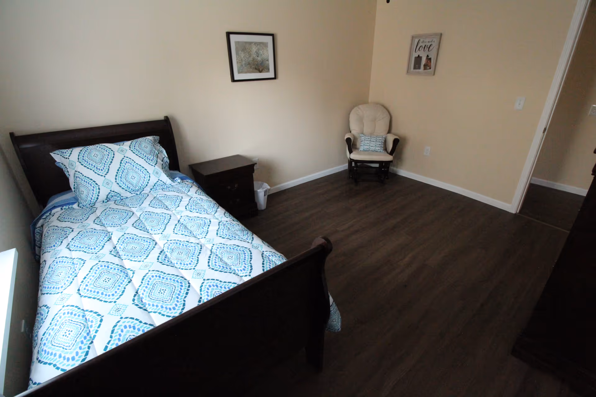 Small furnished bedroom with a twin bed dressed in blue patterned bedding, a nightstand, a cushioned chair in the corner, and dark wood flooring.