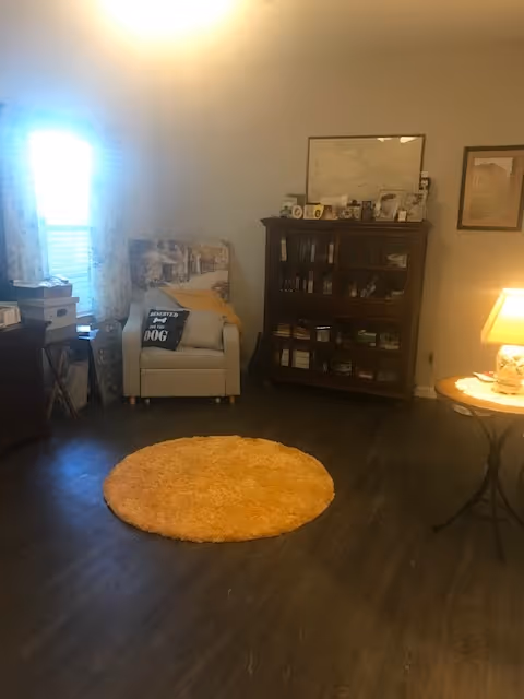 A cozy living room area with a light-colored armchair featuring a pillow that says 'Rescued is my favorite breed'. Next to the chair is a wooden cabinet filled with books and framed photos on top. A round yellow rug is placed on the dark wooden floor. A small round table with a lamp is on the right side, and a window with sheer curtains is on the left.