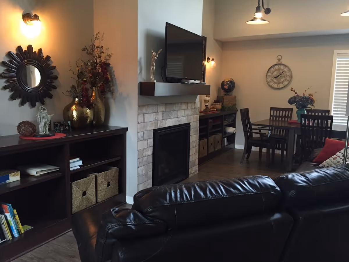 Interior view of a living and dining area in a senior living facility. The foreground shows a black leather couch facing a wall-mounted TV above a fireplace with a stone surround. To the left, there is a dark wooden bookshelf with decorative items including gold vases, a small statue, and a round mirror on the wall above it. In the background, a dining table with four chairs is visible, adorned with a flower arrangement and a large wall clock. The room is softly lit with wall sconces and a hanging light fixture.