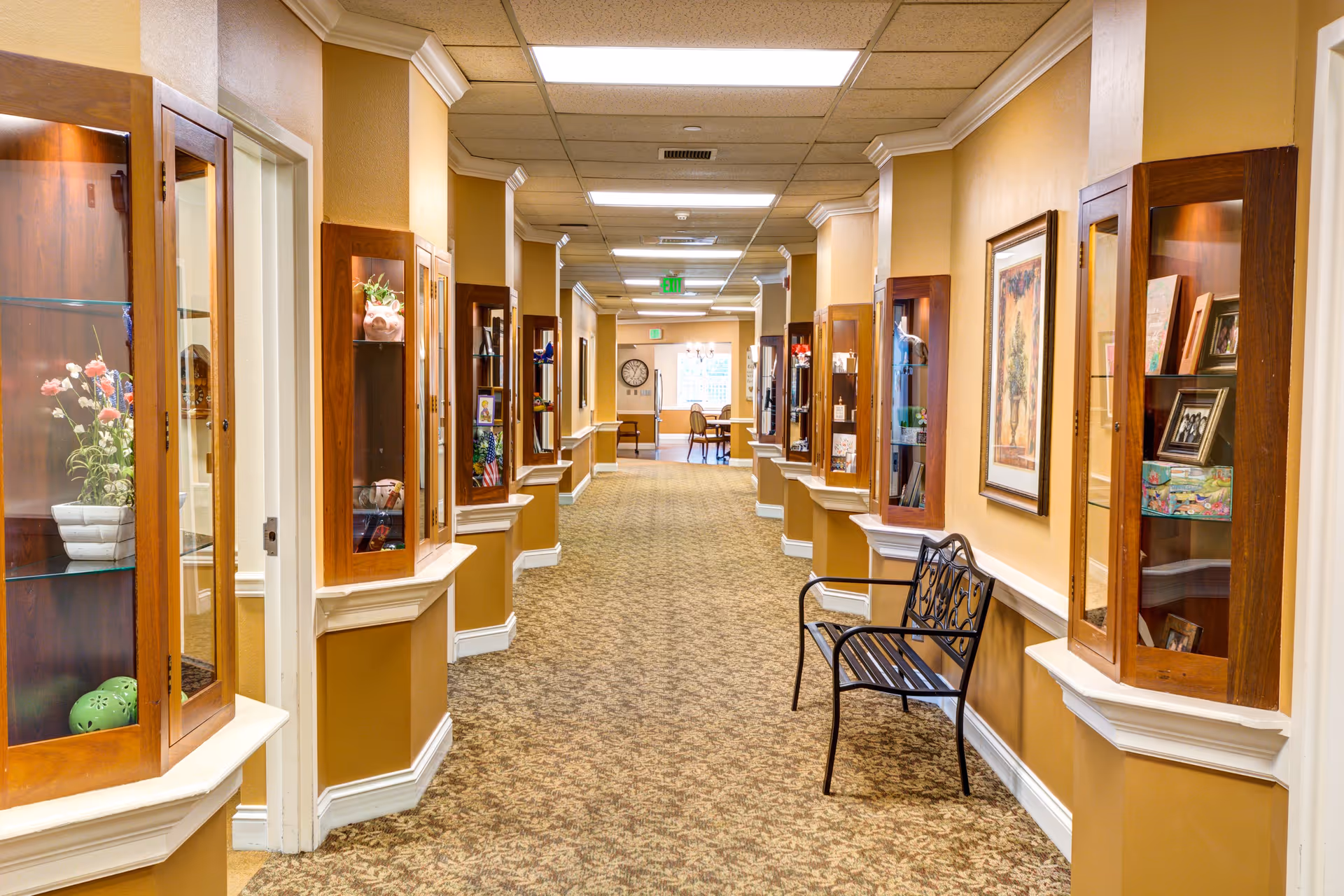 A well-lit hallway in a senior living facility with display cabinets mounted on both walls containing decorative items and framed pictures. The walls are painted in warm tones with white trim, and there is a patterned carpet on the floor. A black metal bench is placed against the right wall, and at the end of the hallway, there is a seating area with chairs and a clock on the wall.