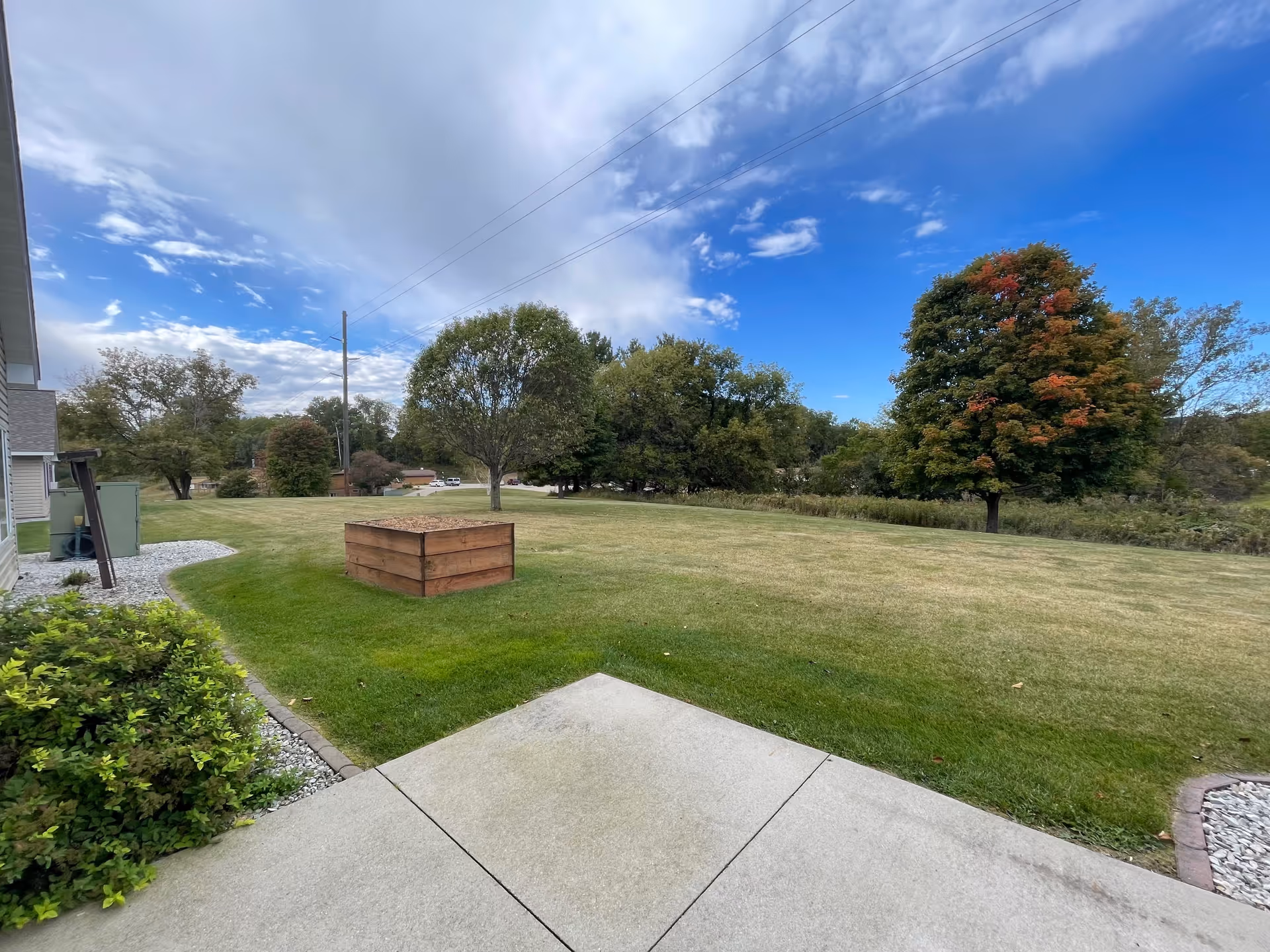 A grassy outdoor area with a few trees, a wooden planter box filled with mulch, and a concrete patio in the foreground. The sky is partly cloudy with patches of blue visible.