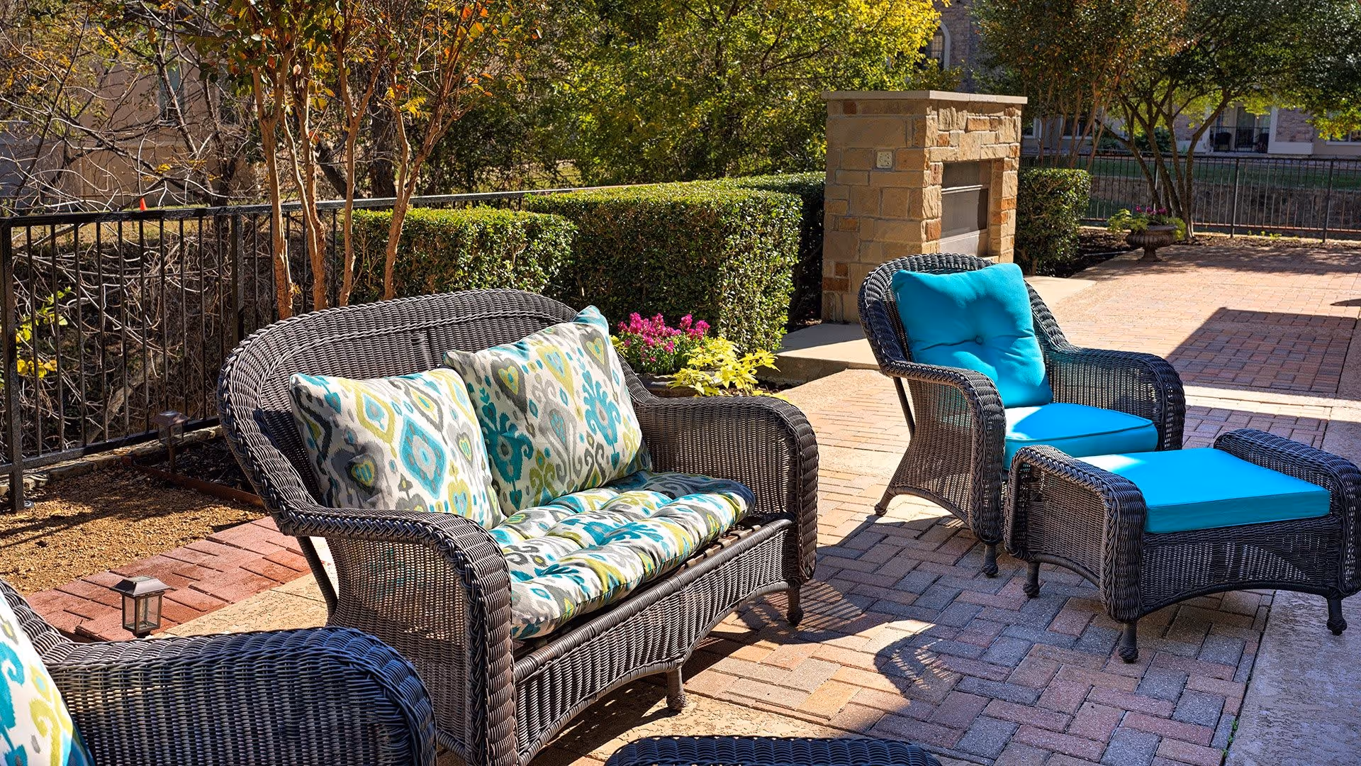 Outdoor patio area with wicker furniture including a loveseat with patterned cushions, a chair with bright blue cushions, and a matching ottoman. The patio is paved with bricks and features a stone fireplace in the background, surrounded by greenery and trees.