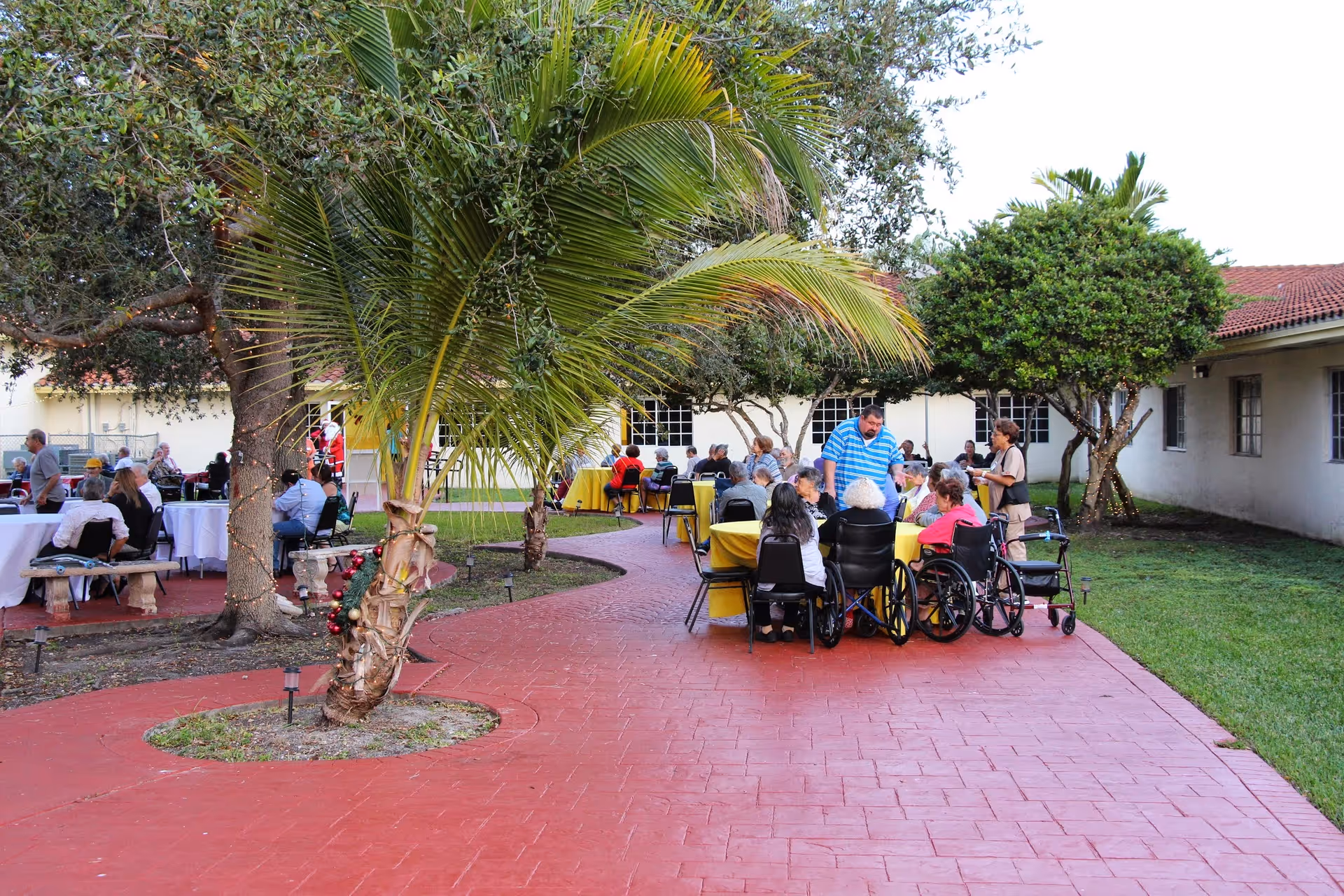 Outdoor patio area at Floridian Gardens Assisted Living Facility with elderly residents sitting at tables covered with yellow and white tablecloths. Some residents are in wheelchairs, and a few staff members are attending to them. The area is surrounded by trees and greenery, with a red paved walkway.