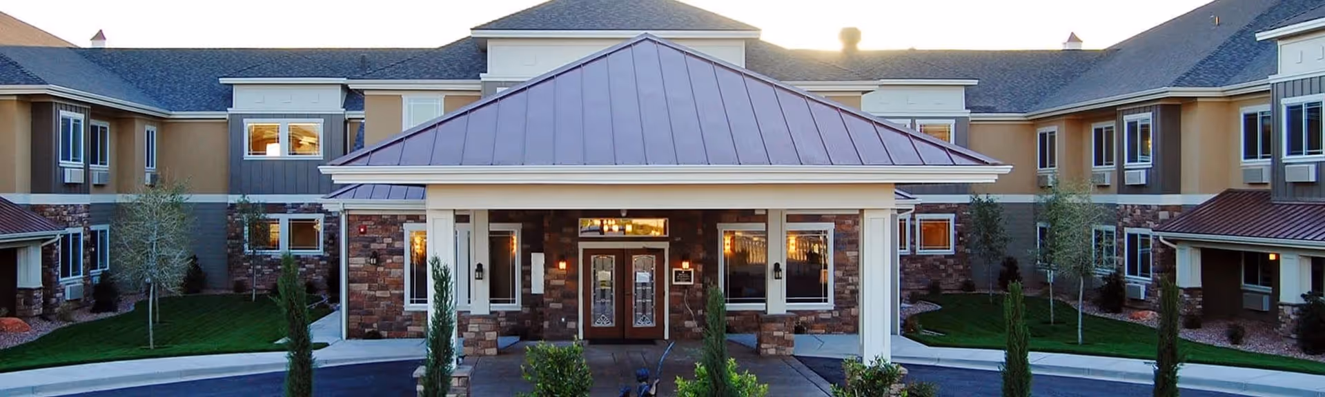 Front exterior view of a senior living facility named Spring Gardens St. George, featuring a covered entrance with double glass doors, stone and beige siding, multiple windows, and a landscaped driveway with small trees and shrubs.