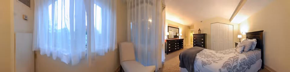 A bright bedroom with a single bed featuring a patterned gray and white bedspread and pillows. The room has large windows with sheer white curtains allowing natural light to fill the space. There is a white chair near the windows, a dark wooden dresser with a mirror, and another tall dresser. The walls are painted a light color, and the room has a cozy, clean atmosphere.