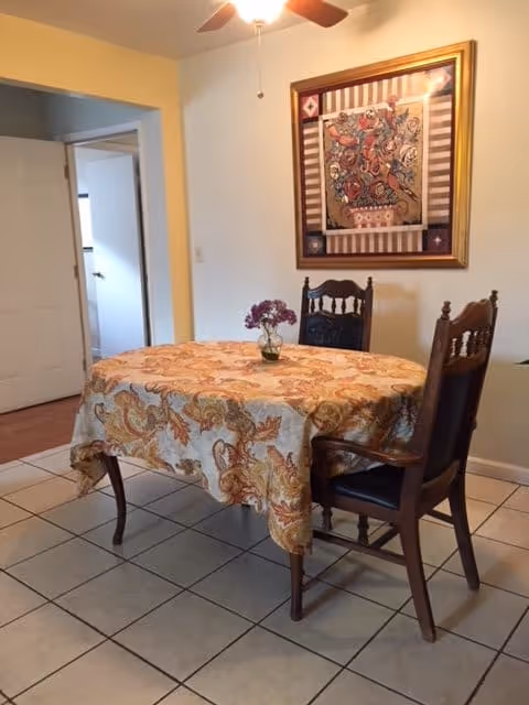 A small dining area with a rectangular table covered with a patterned tablecloth in shades of orange and beige. Two wooden chairs with dark cushions are placed at the table. A small vase with purple flowers is centered on the table. On the wall behind the table, there is a framed artwork featuring a floral design. The floor is tiled in white, and a ceiling fan with a light is visible above.
