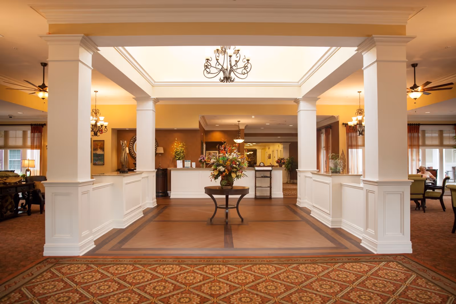 Spacious and elegant reception area of a retirement community featuring a central table with a large floral arrangement, white columns, chandeliers, warm lighting, and comfortable seating areas on both sides.