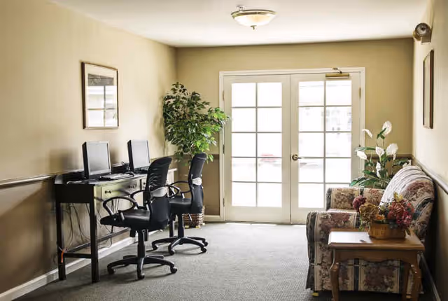 A small seating and computer area in a senior living facility with two black office chairs at a desk with two desktop computers on the left, a floral-patterned couch and a wooden side table with a flower arrangement on the right, a large potted plant near the center, and double glass doors letting in natural light at the back.