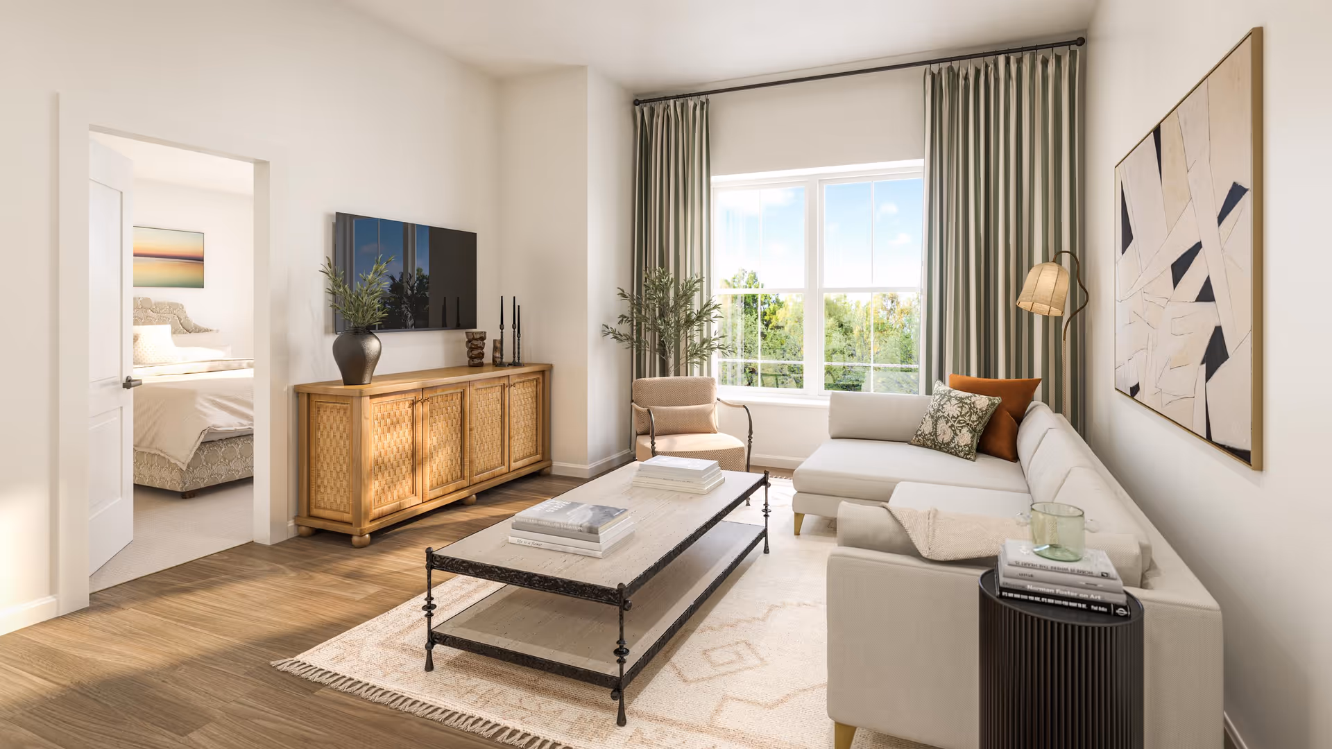 Bright living room with an L-shaped beige sectional sofa, a coffee table, a wooden media console under a wall-mounted TV, and a large window with curtains.