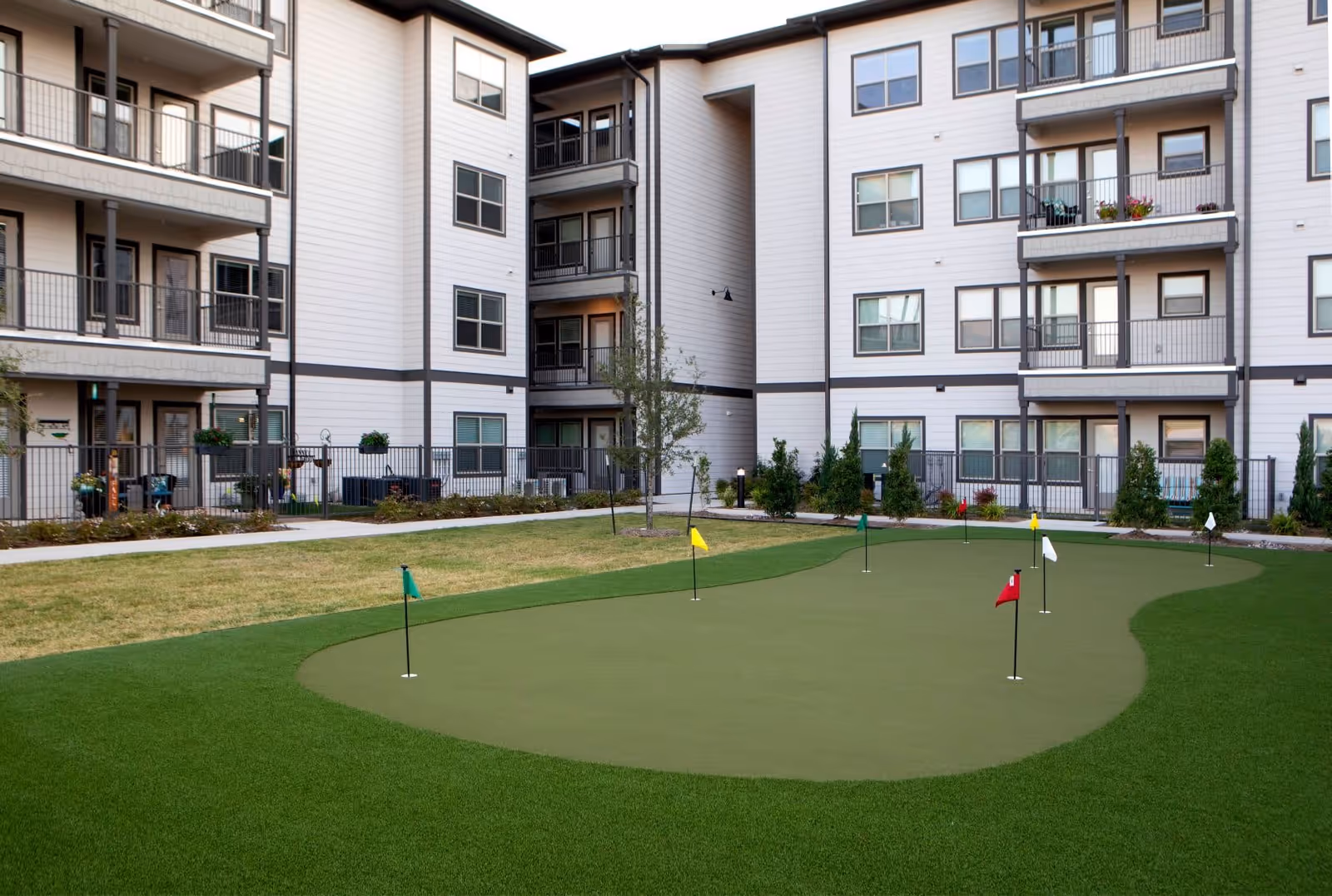 Outdoor putting green with multiple small flags in front of a multi-story residential building with balconies and windows.