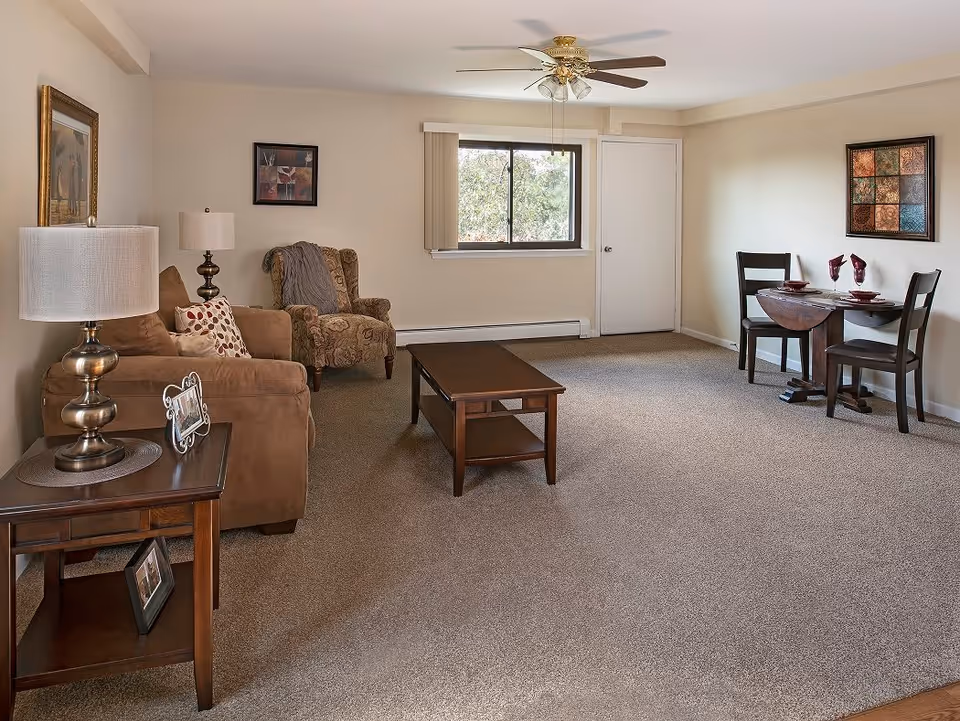 A living room with beige walls and carpeted floor featuring a brown sofa with patterned cushions, a patterned armchair, a wooden coffee table, and a wooden side table with a lamp and picture frames. There is a ceiling fan with lights, a window with vertical blinds, and a small dining area with a round wooden table set for two and two chairs. Two framed artworks hang on the walls.