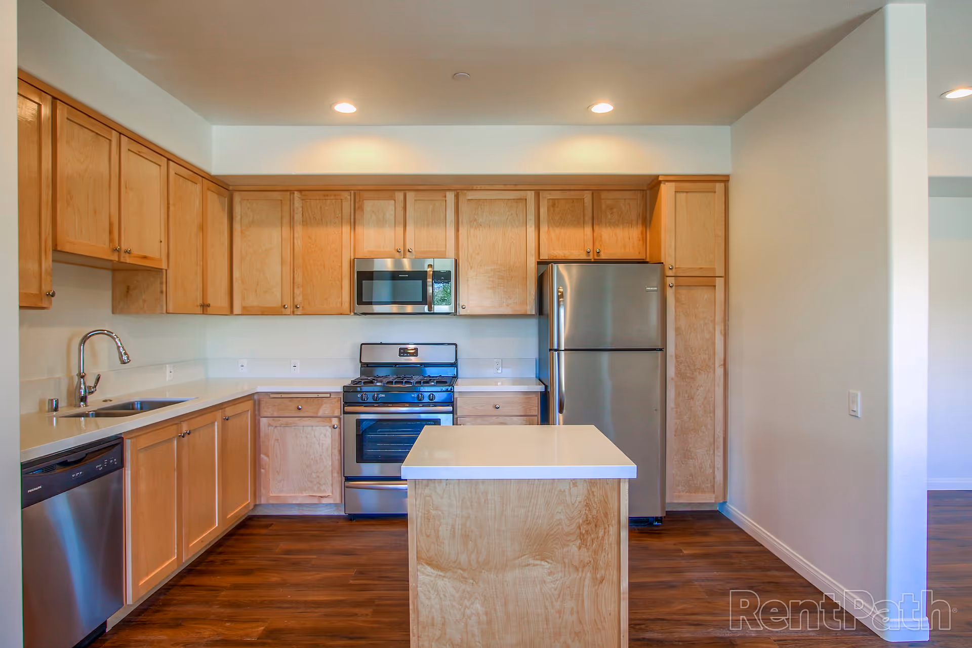 Modern kitchen with light wood cabinets, stainless steel appliances including a refrigerator, microwave, stove, and dishwasher, a white countertop island in the center, and recessed ceiling lights.