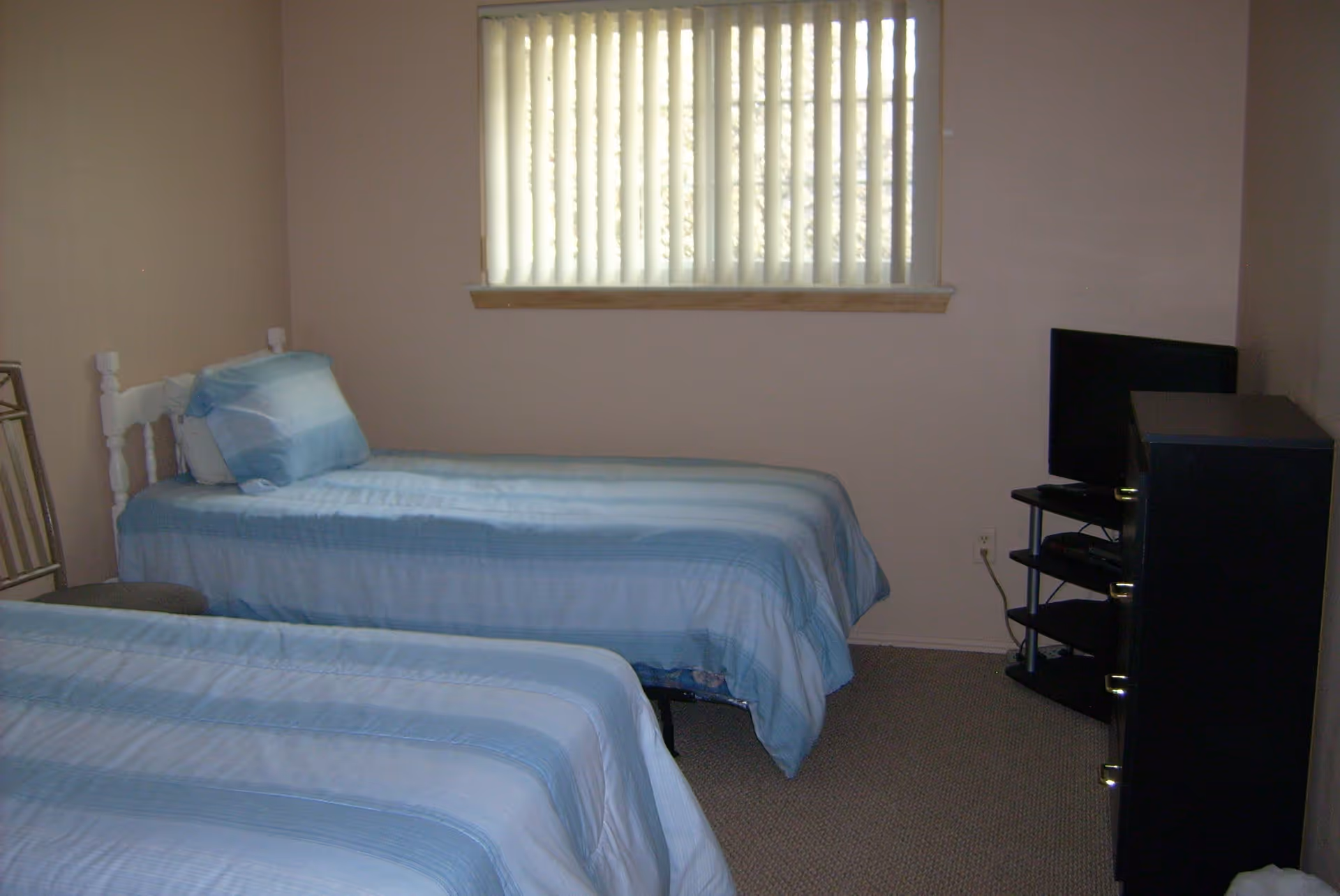 A simple bedroom with two twin beds covered in blue striped bedding. There is a window with vertical blinds on the wall behind one bed. A small black TV stand with a flat-screen TV is positioned in the corner of the room. The walls are painted light beige and the floor is carpeted.