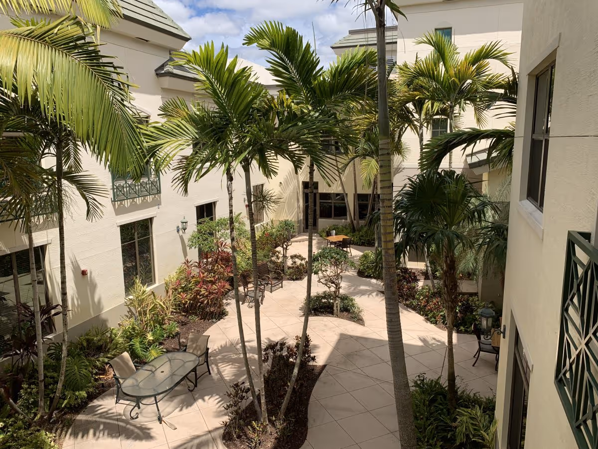Outdoor courtyard area at Luxe Senior Living at Wellington featuring tall palm trees, various plants, a tiled walkway, and outdoor seating with tables and chairs surrounded by light-colored building walls.