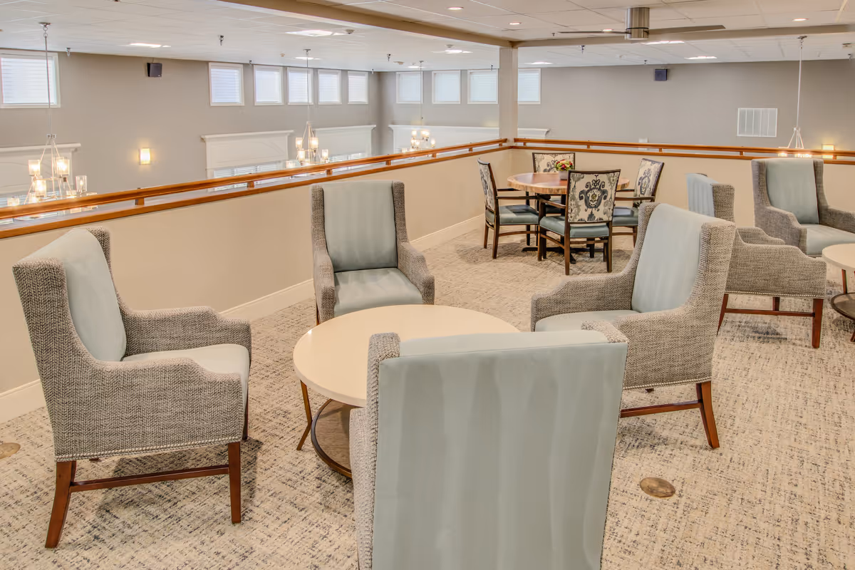 A bright communal lounge with upholstered armchairs arranged around round tables overlooking a lower-level atrium.