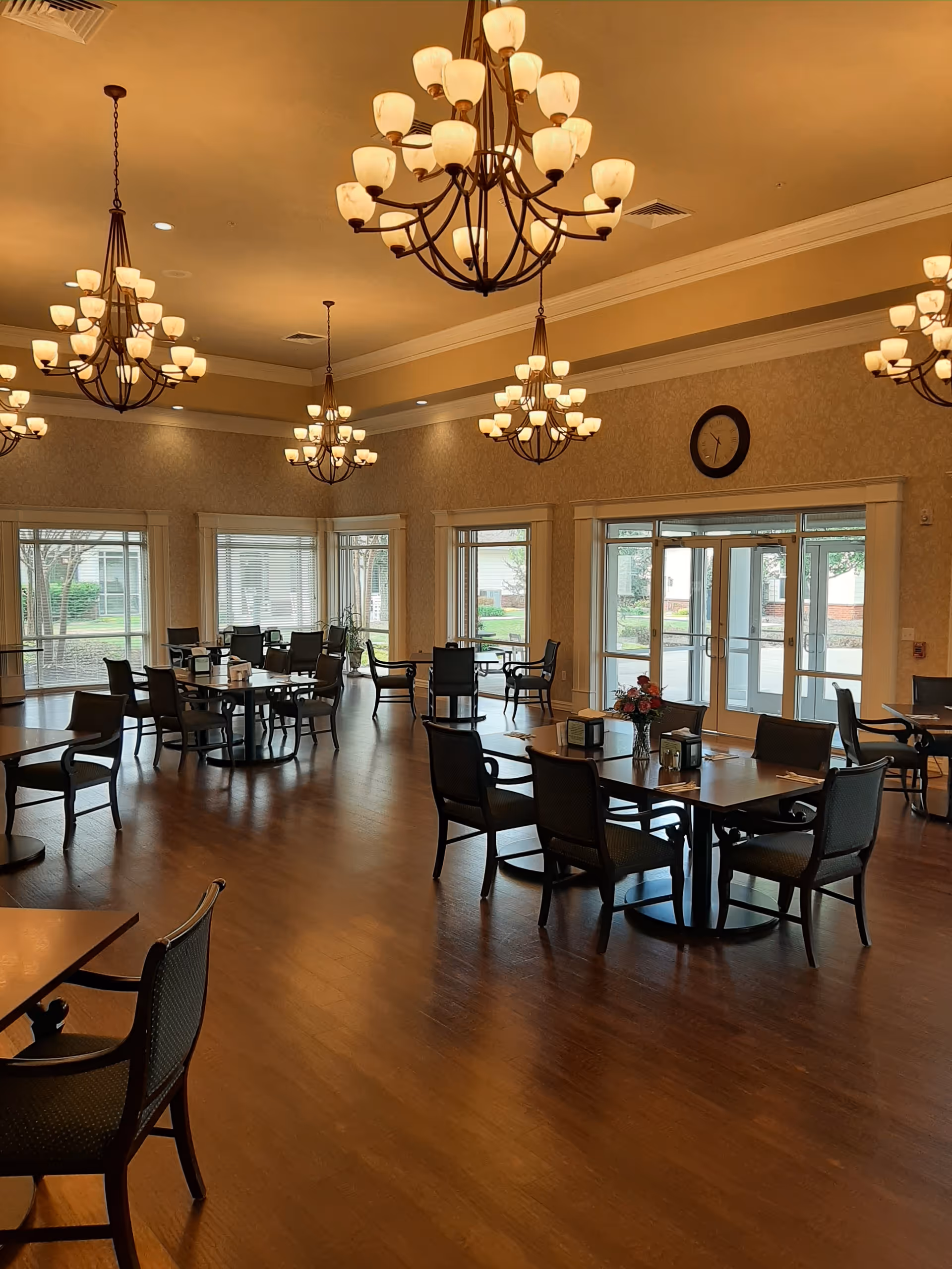 A spacious dining room with multiple round tables and chairs arranged neatly. The room features large windows allowing natural light to enter, elegant chandeliers hanging from the ceiling, and a clock on the wall above the entrance doors.