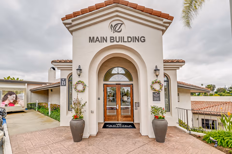Entrance to the main building of a senior living facility with a beige stucco exterior, arched doorway, wooden double doors, two large potted plants with flowers on either side, and decorative wreaths hanging on wall-mounted lanterns. The building has a tiled roof and a sign above the entrance that reads 'MAIN BUILDING'.