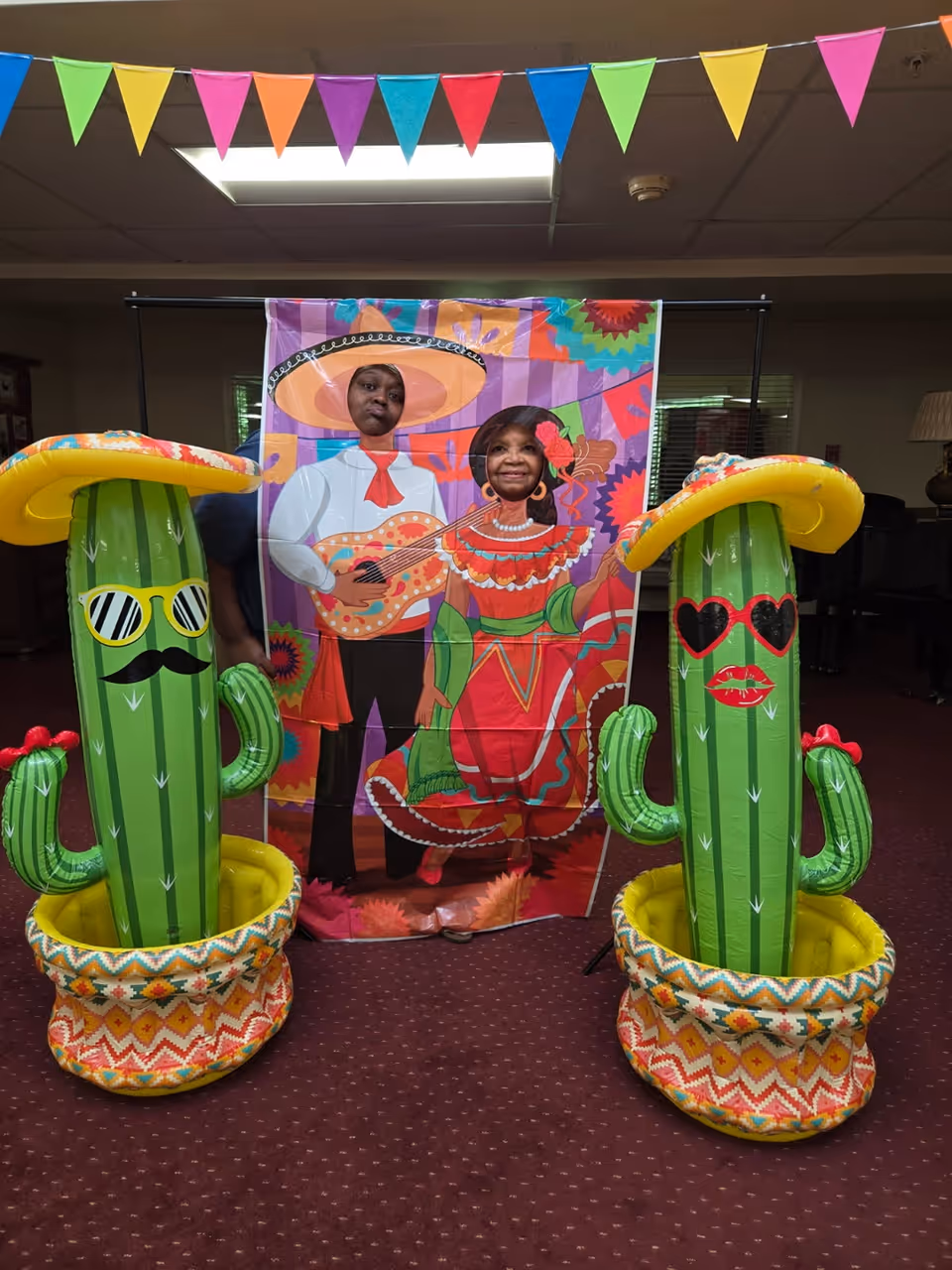 Two people posing behind a colorful cutout standee depicting a man playing guitar and a woman in traditional Mexican attire. They are flanked by two inflatable cacti wearing sombreros and sunglasses, with one cactus having a mustache and the other red lips. Colorful triangular pennant banners hang above them in an indoor setting with carpeted floor.
