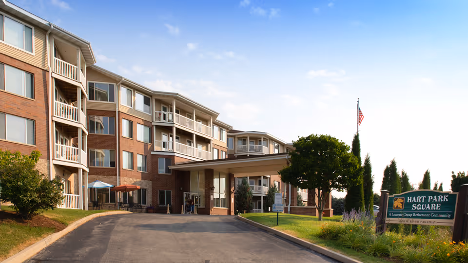 Front exterior of a multi-story brick senior living building with a covered entrance, driveway, landscaping, and a Hart Park Square sign.
