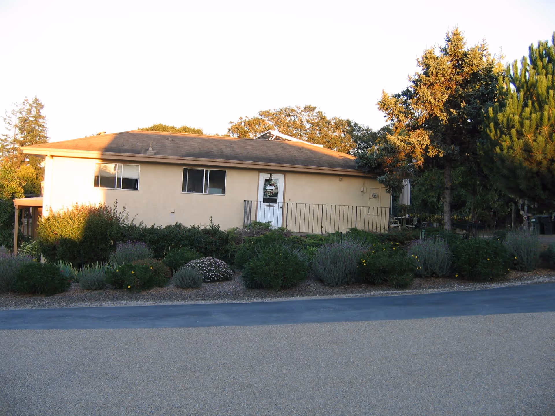 Exterior view of a single-story residential building with beige walls and a brown roof, surrounded by green shrubs and trees, under a clear sky during daylight.