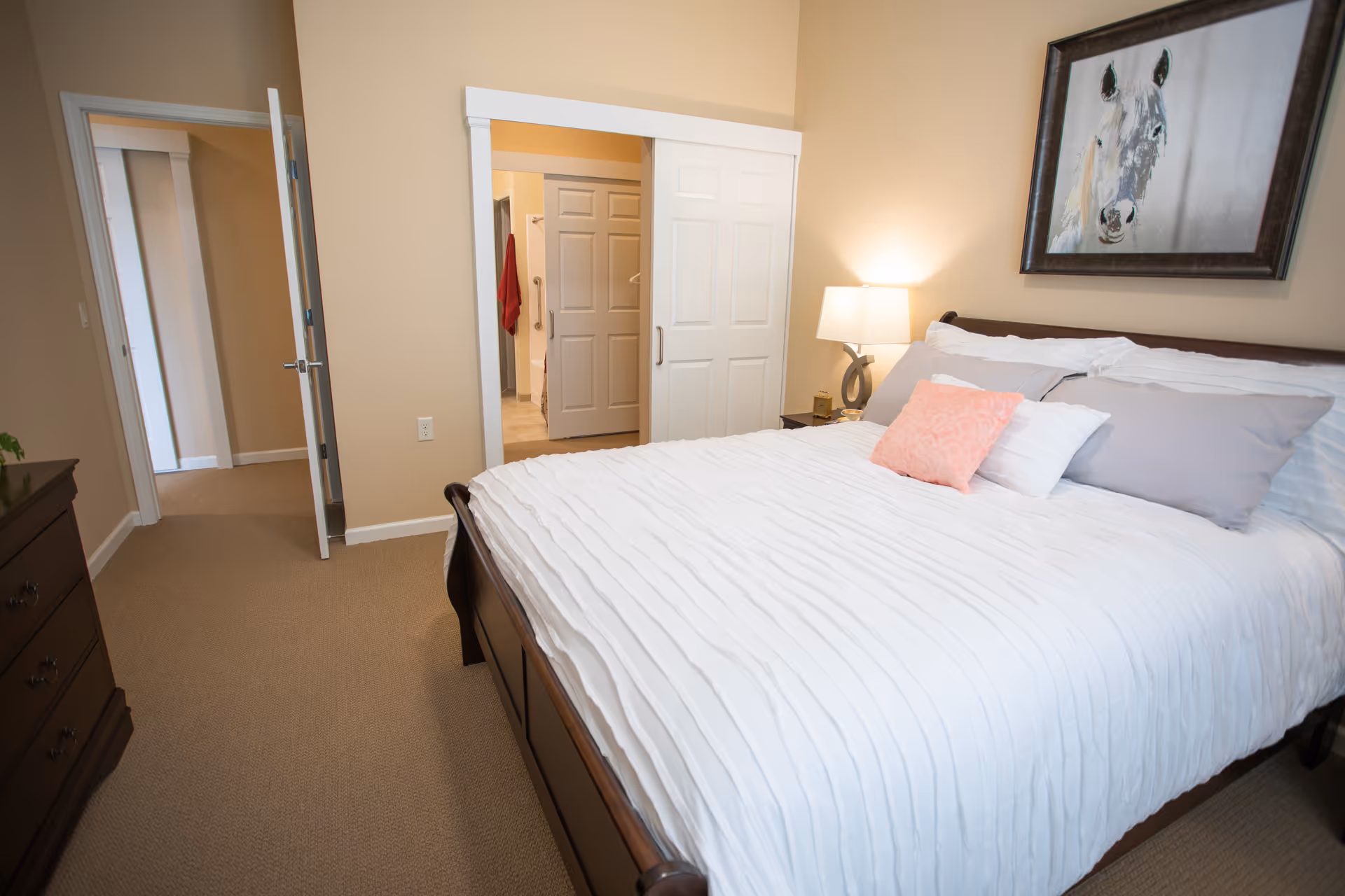 A neatly made bed with white bedding and several pillows, including a pink decorative pillow, in a bedroom with beige walls. A nightstand with a lamp is beside the bed, and a framed painting of a horse hangs above the headboard. There is an open door leading to a hallway and a partially open closet door revealing a bathroom with a red towel hanging inside.