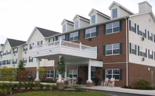Exterior view of a multi-story assisted living facility with a combination of brick and siding facade, featuring a covered entrance with white columns and a balcony above. There are several windows, some with dormers, and a landscaped area with small trees and shrubs in front.