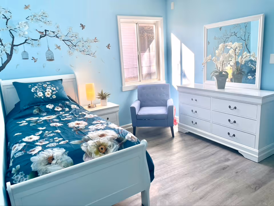 A bright bedroom with light blue walls featuring a floral and bird mural. The room contains a white single bed with a blue floral bedspread, a white nightstand with a small lamp and plant, a blue armchair near a window, and a white dresser with a large mirror and a vase of white flowers on top.