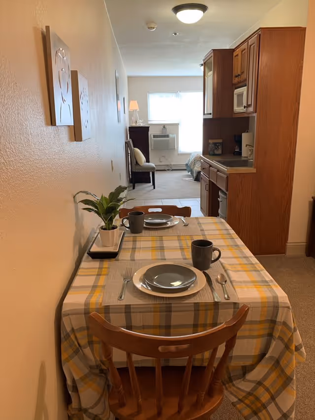 A small dining area with a wooden table covered by a yellow and gray plaid tablecloth, set for two with plates, mugs, and cutlery. A small potted plant is on the table. The dining area is adjacent to a compact kitchen with wooden cabinets and a microwave. In the background, there is a chair, a lamp, a window with blinds, and a bed partially visible, suggesting a studio apartment layout.