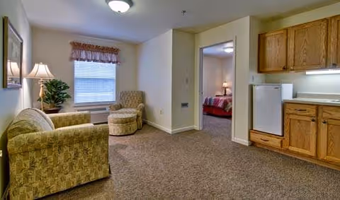 A cozy living area with a patterned armchair and matching ottoman, a side table with a lamp and a plant, a window with a valance, beige walls, and carpeted floor. To the right, there is a kitchenette with wooden cabinets, a small refrigerator, and a countertop. An open doorway leads to a bedroom with a bed and a lamp visible.