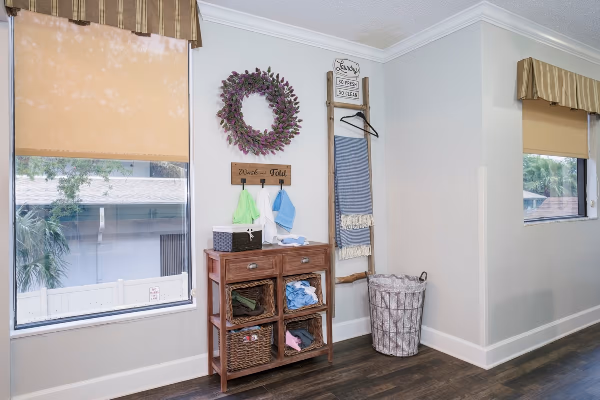 A bright laundry area with two windows covered by beige roller shades and striped valances. A wooden cabinet with four wicker baskets holds folded clothes and laundry items. Above the cabinet is a decorative wreath and a sign that reads 'Wash and Fold.' Next to the cabinet is a wooden ladder with a hanging blue towel and a sign that says 'Laundry So Fresh So Clean.' A large laundry basket is placed on the floor beside the ladder.