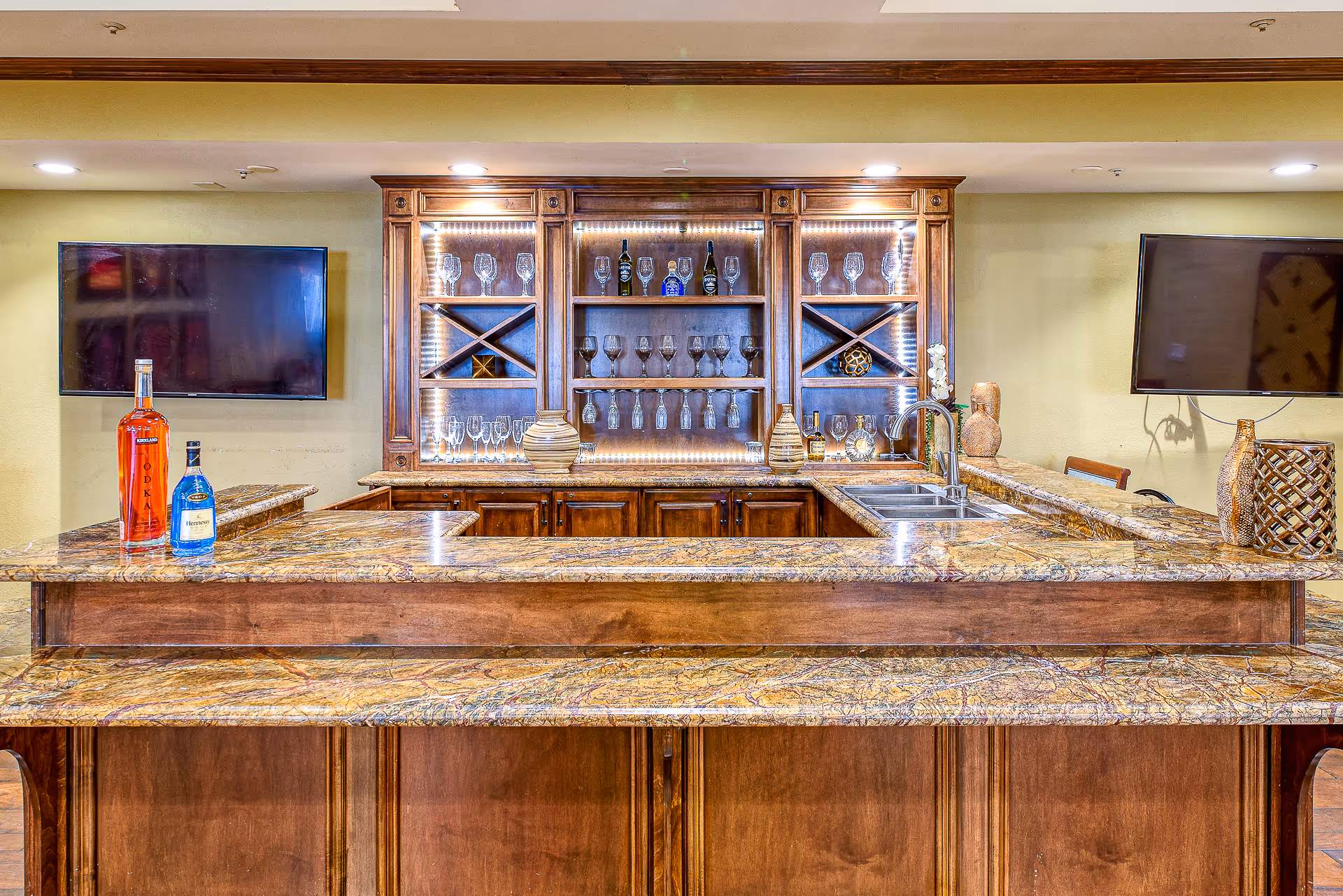 A well-lit bar area with a marble countertop and wooden cabinetry. The back wall features shelves with various wine glasses and bottles, illuminated by LED lights. Two flat-screen TVs are mounted on the wall on either side of the bar. Decorative vases and bottles are placed on the counter and shelves.