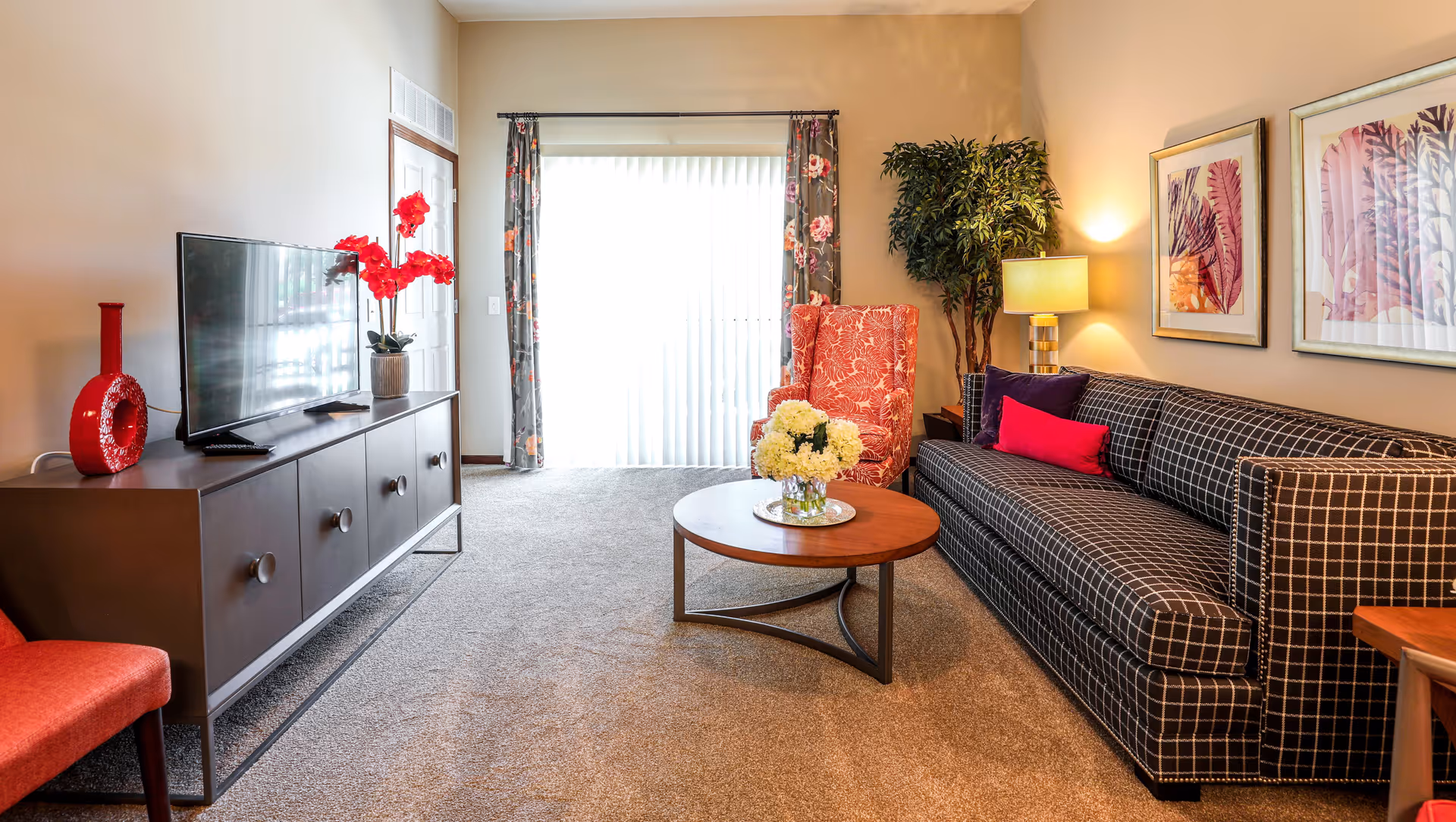 A cozy living room with a checkered black and white sofa adorned with colorful pillows, a round wooden coffee table with a vase of white flowers, a red patterned armchair, a TV on a dark wooden cabinet, and a tall green plant next to a lit table lamp. The room has beige walls, carpeted floor, floral curtains, and framed artwork on the wall.