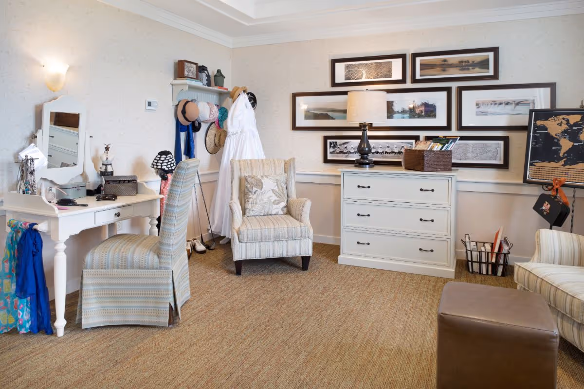 Cozy interior room with upholstered chairs, a vanity desk, a dresser with a lamp, and framed wall art on the wall.