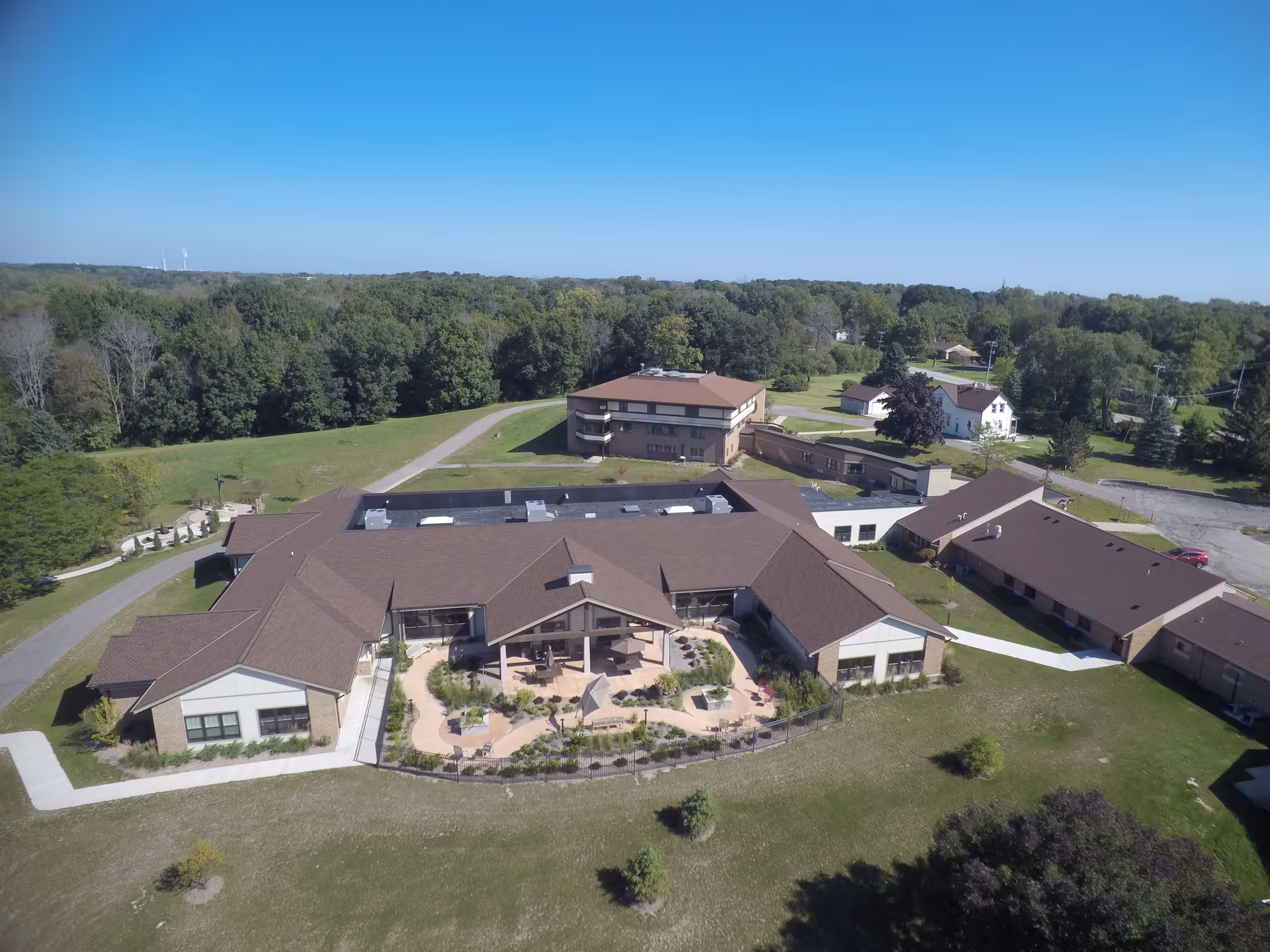 Aerial view of St Monica's Senior Living facility showing multiple connected buildings with brown roofs surrounded by green lawns and trees under a clear blue sky.