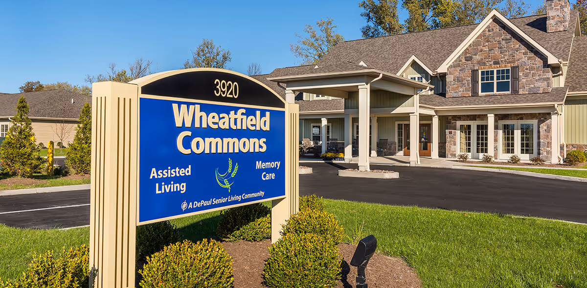 Exterior view of Wheatfield Commons, a senior living community building with a stone and siding facade, a covered entrance, and a blue sign in the foreground displaying the name, address 3920, and services including Assisted Living and Memory Care.